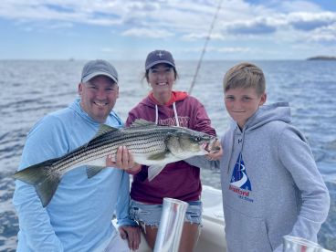 A family proudly displaying a large Striped Bass while fishing on a boat, enjoying a day on the water.