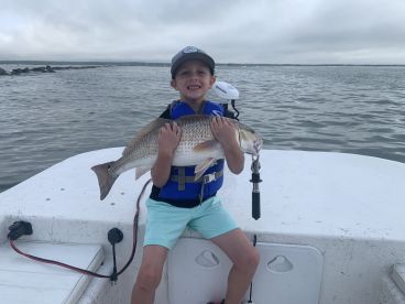 A young angler proudly displaying a large Redfish while on a boat, showcasing a successful fishing trip.