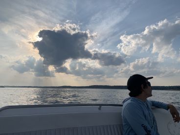 A lone angler enjoys the serene view from the boat, with dramatic clouds and sunlight reflecting on the water.