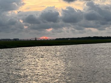A scenic view of a sunset over the water, with a distant bridge and boat visible, capturing the tranquil atmosphere of a fishing environment.