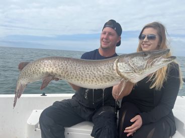 Two anglers proudly displaying a large Muskellunge while fishing on a boat, showcasing a successful day on the water.