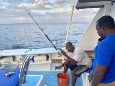 Two men and a child are aboard a fishing boat, with one man preparing gear while the other observes. The calm sea and cloudy sky create a serene fishing atmosphere.