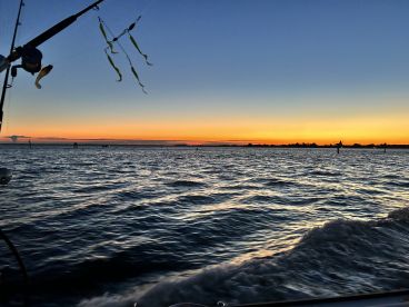 A serene sunset view from the side of a fishing boat, showcasing the calm waters and distant silhouettes of other boats on the horizon.