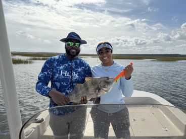 Two anglers proudly displaying a large Black Drum while fishing in a scenic coastal environment.
