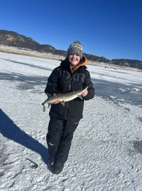 A young angler proudly displaying a Northern Pike caught while ice fishing in a scenic mountain lake.
