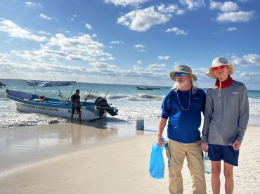 A father and son stand on the beach next to a fishing boat, ready for a day of fishing in the clear waters of Tulum.