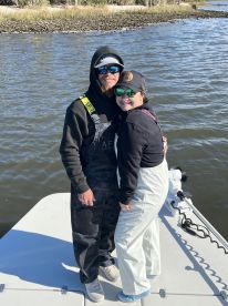 A couple posing on the bow of a fishing boat, dressed in fishing gear, with a scenic waterway in the background.