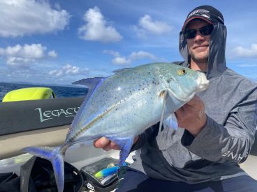 An angler proudly displaying a vibrant Bluefin Trevally while fishing on a boat in clear blue waters.