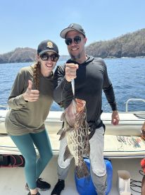 Two anglers proudly displaying a large Leopard Grouper while fishing on a sunny day in a coastal environment.