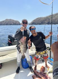 Two anglers proudly displaying a large Grouper caught during a deep-sea fishing trip, with several other fish visible on the deck.