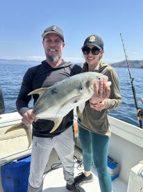 Two anglers proudly displaying a large Yellowtail while fishing on a boat in a sunny coastal environment.