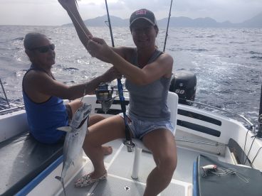 Two anglers aboard a boat, one proudly holding a freshly caught fish while the other assists, set against a scenic ocean backdrop.