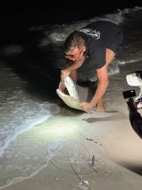Angler reeling in a shark at night on the beach, showcasing the thrill of surf fishing.