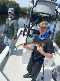 A young angler proudly displaying a Snook caught while fishing in a mangrove area, with a guide nearby.