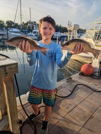 A young angler proudly displaying two Snapper catches at the dock, showcasing a successful fishing trip.