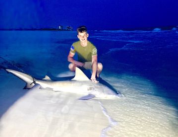 A young angler proudly displaying a caught shark on the beach at night, showcasing the thrill of surf fishing.