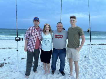 A family group poses on the beach with fishing rods in the background, enjoying a day by the ocean.