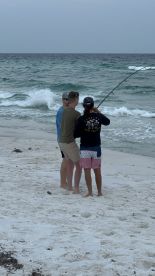 Three young anglers fishing on the beach, with one actively reeling in a line while the others watch attentively.