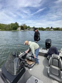 Two anglers actively fishing from a Lund boat on a serene lake, with one preparing to net a catch while the other casts a line.