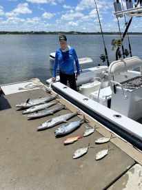 An angler stands proudly beside a well-arranged catch of various fish, including King Mackerel and Snapper, displayed on the dock after a successful fishing trip.