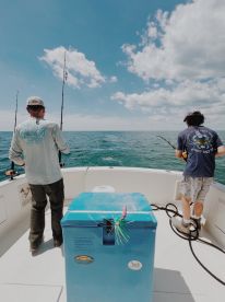 Two anglers actively fishing on a boat, casting lines into the open water under a bright blue sky.