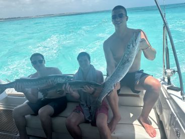 Three young anglers proudly displaying their catch of Barracuda while enjoying a sunny day on a boat in clear turquoise waters.