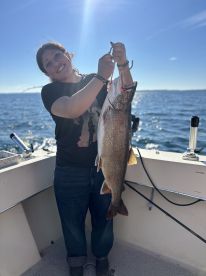 An excited angler proudly displaying a large Lake Trout while fishing on a boat, with a clear blue sky and water in the background.