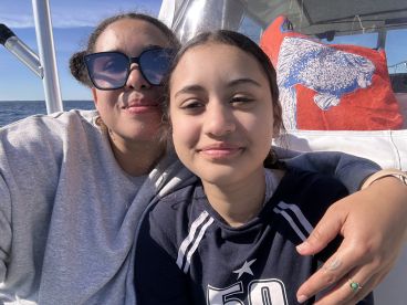 Two young anglers enjoying a sunny day on a boat, with a scenic ocean backdrop and a playful fish-themed bag in view.