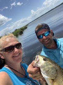 Two anglers smiling while holding a large Speckled Trout in a scenic waterway, showcasing a successful day of fishing.
