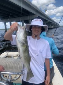 A young angler proudly displaying a striped bass while fishing under a bridge, showcasing a sunny day on the water.