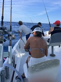 A group of anglers actively fishing on a boat, with a young girl in a bikini and shorts focused on reeling in a catch in open waters.