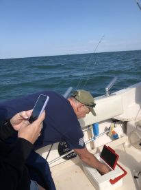 A fisherman is seen inside the boat, checking a cooler while another person captures the moment on their phone, showcasing a casual fishing experience.