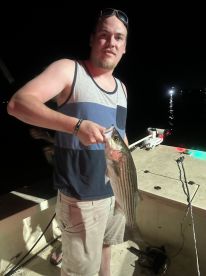 Angler holding a striped bass while fishing at night, showcasing a successful catch on a boat.