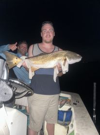 Angler proudly displaying a large Redfish while on a night fishing trip, with a fellow fisherman giving a thumbs up in the background.