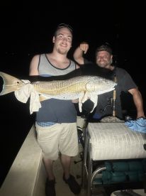 Two anglers proudly displaying a large Redfish aboard a boat during a nighttime fishing trip.