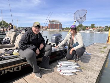 Two anglers posing with a catch of multiple fish on the dock after a successful fishing trip, with a scenic marina backdrop.