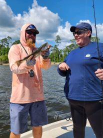 Two anglers on a boat enjoying a successful fishing trip, showcasing a Speckled Trout against a scenic backdrop.