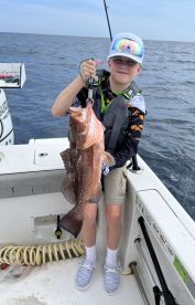 A young angler proudly displaying a large Grouper while fishing on a boat in open waters.