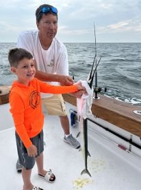 A young boy joyfully holds a large fish, likely a Mackerel, with the help of an adult on a fishing boat, showcasing a memorable moment on the water.