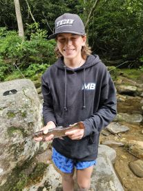 Young angler proudly holding a small trout while standing by a stream, showcasing a successful catch in a natural setting.