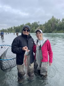 Two anglers proudly displaying their Silver Salmon catch while wading in a river, surrounded by a scenic backdrop of trees.