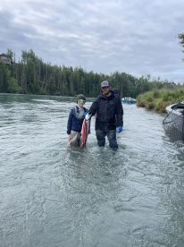 A young angler and an adult proudly displaying a large salmon while wading in a river, showcasing a successful fishing trip.