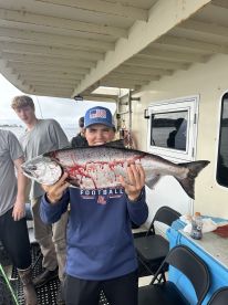 Young angler proudly displaying a large Salmon aboard a fishing charter, surrounded by fellow anglers.