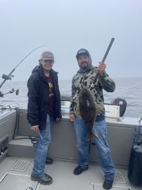 A young angler and an adult proudly display a large Halibut aboard a fishing boat, showcasing their successful catch during a foggy day on the water.