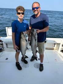 A young angler and an adult proudly displaying their catch of two large fish while on a fishing charter in open waters.