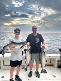 A young angler and an adult proudly displaying their catch of two large salmon while fishing on a boat, with a scenic ocean backdrop.