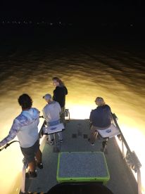 Group of anglers fishing at night from a boat, illuminated by the vessel's lights, creating a vibrant atmosphere on the water.