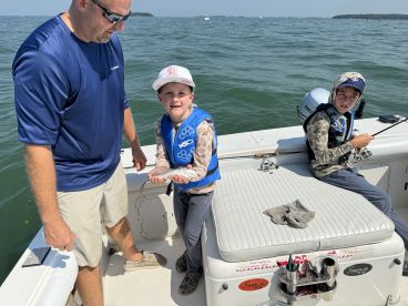 A young angler proudly displaying a caught fish while fishing on a boat, accompanied by an adult and another child, enjoying a day on the water.