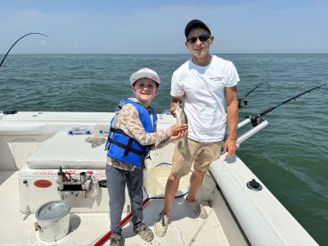 A young angler in a life jacket proudly displays a caught fish alongside an adult on a fishing charter in open waters.