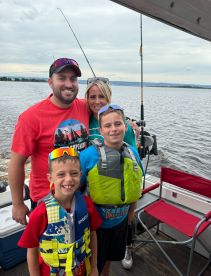 A family enjoying a fishing trip on a boat, with two boys in life jackets and their parents smiling in the background.